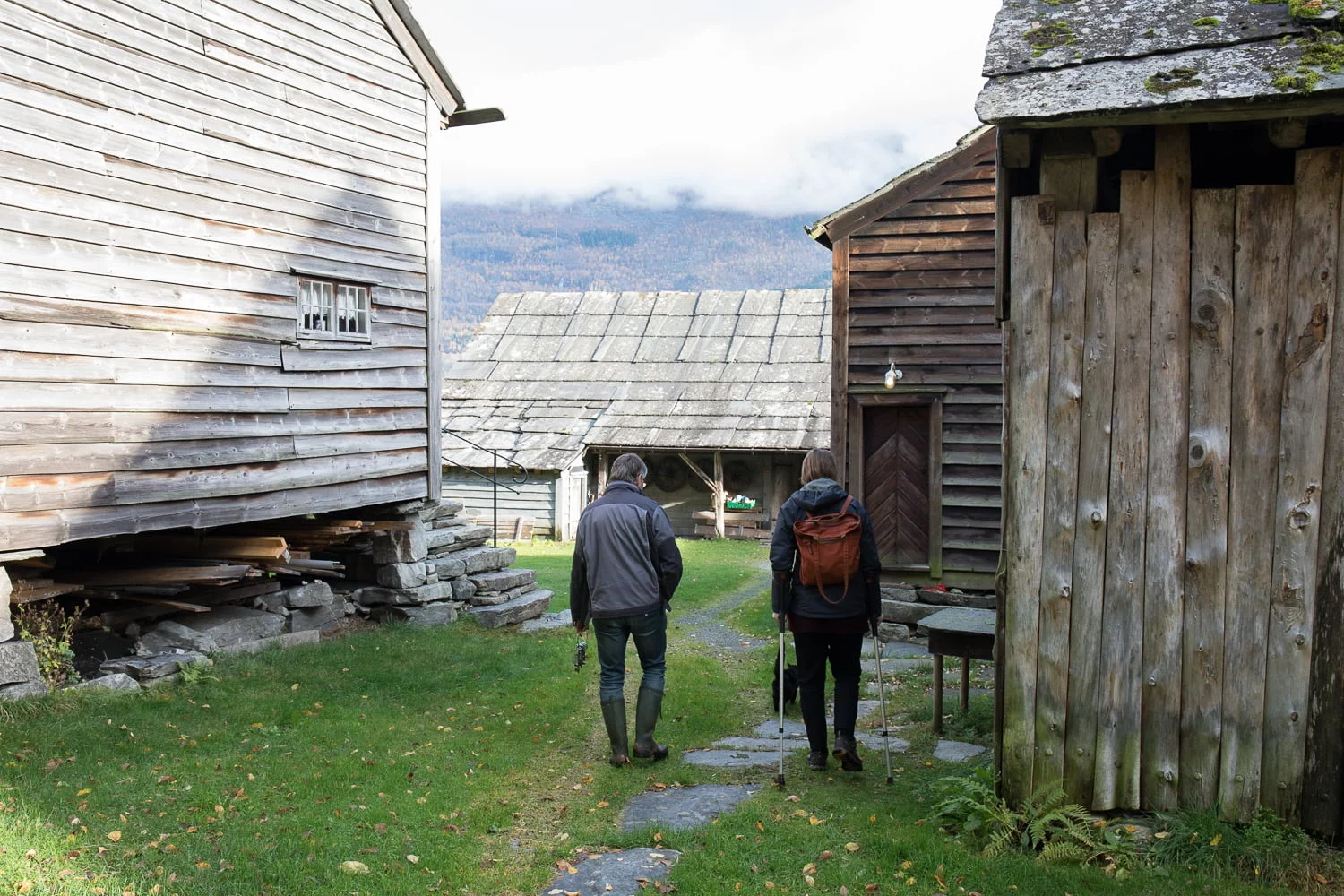 To menn sett bakfrå gjeng mellom gamle, umåla bygninger. Ein av dei nyttar to krykker. Dei føl ein sti av trakkheller over graset. Bak husa ser ein høge fjell. Husa har skifertak med store heller.