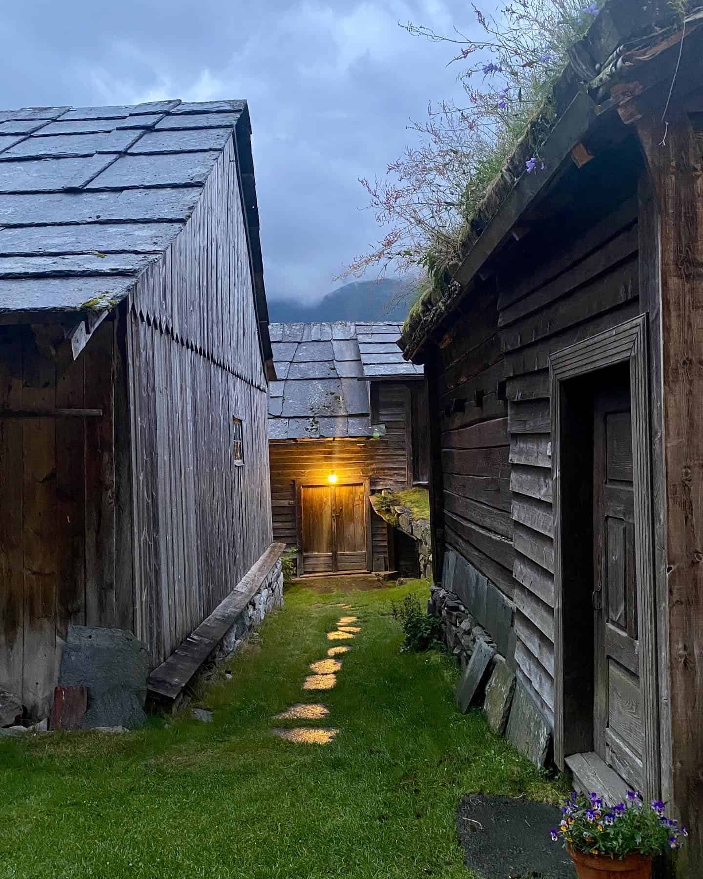 Ei skrånande veg med trakkheller mellom gamle, umåla bygninger - ein med helletak og ein med torvtak, ned mot ei gamal løe. Det er skumring, og litt snø på fjella bak løa. Stemningsfullt ljos fra utelampa på løo reflekterar i trakkhellene, dei skin. 