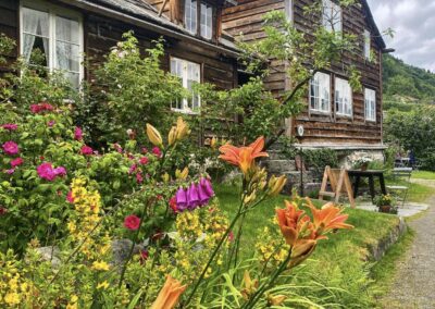 Eit gamalt hus med skifertak og tjærebreidd panel ses langs ein grusveg og mot ei trekledt fjellside. Vindauga er kvitmåla og har blondegardiner. I forgrunnen eit vell av fargerike blomer - gul fredlaus, oransje liljer, lilla revebjølle, rosa nyperoser, ein grasflekk og eit lite frukttre. Utanfor steintrappa opp til døra står eit bord med ei kvit potteplante. Frodig og stemingsfull plass!