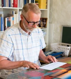 Ein mann med briller. grånande hår og velfrisert grått skjegg i kvit, korterme skjorte sit ved eit bord og gjer klar eit papir til kalligrafi. Bak han velfylte bokhyller. Han smiler lett og ser konsentrert ut.