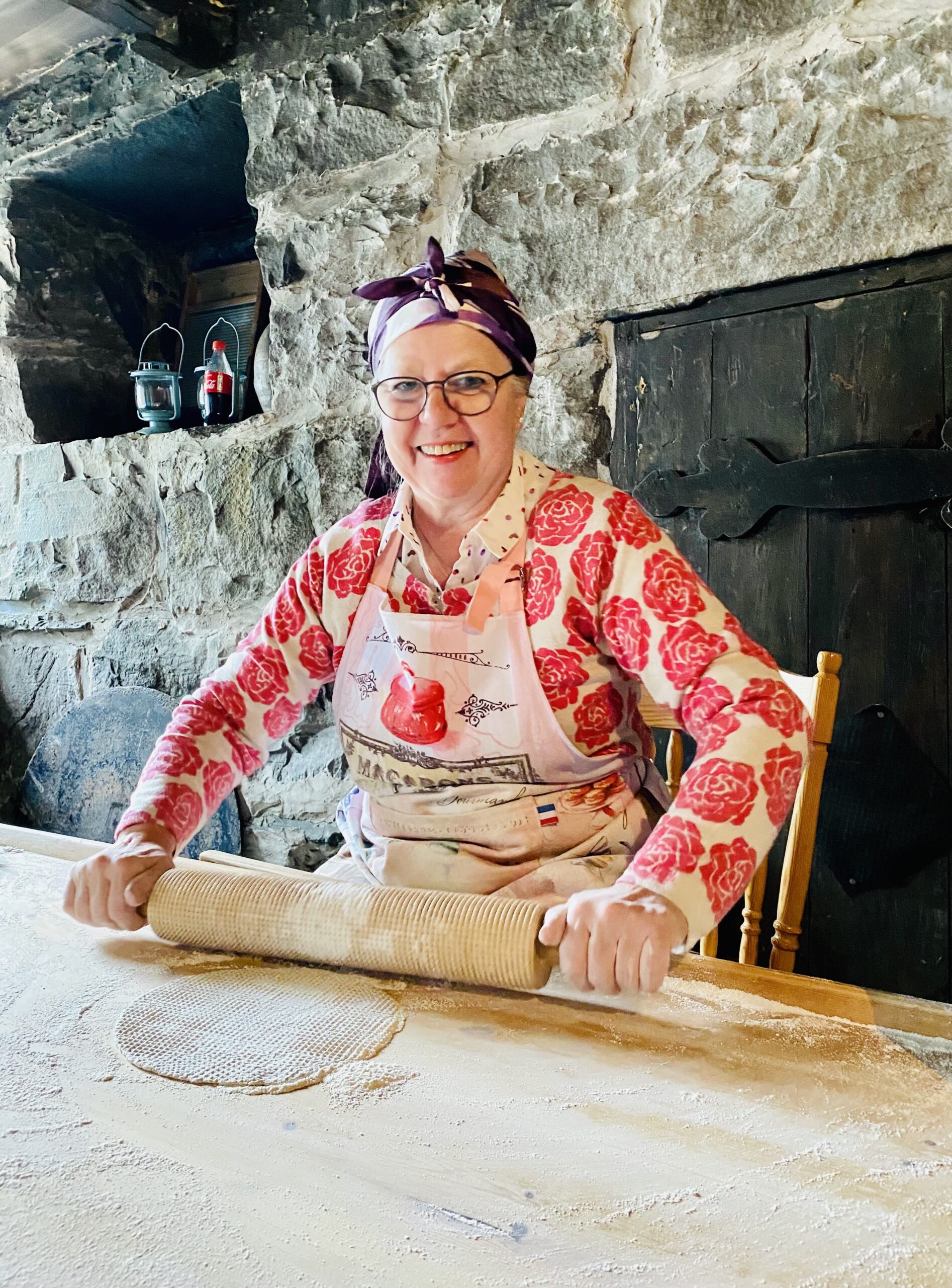 Ei blid dame sit ved eit bakstebord i en steinkjeller. Ho har eit kjevle i nevane og ein leiv på bordet. Ho har briller, eit silkesjal knytt på hovudet og ein kvit cardigan med mønster av store raudrosa roser, og ein kvitt forkle.