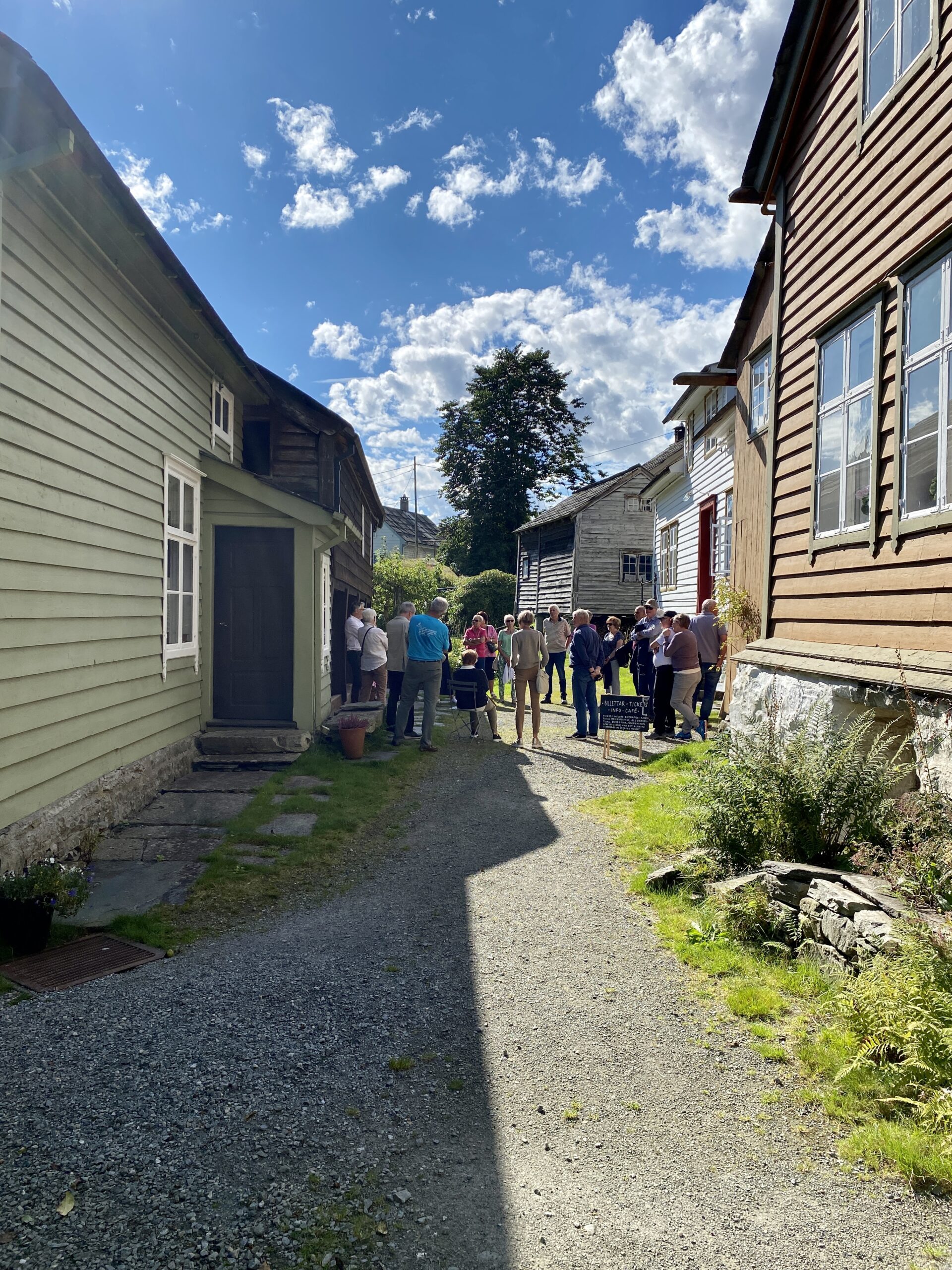 Ei brei grusveg mellom gamle hus. mot enden av vegen ei gruppe mennesker som lyttar til ein omvisar. Det er ein fin solskinnsdag, og skygge fra husa til venstre ligg over halve vegen.